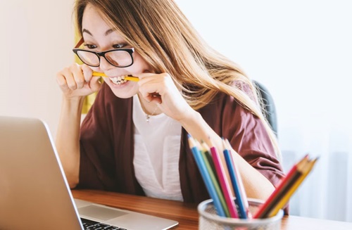 A university student wearing glasses sits at a desk, biting a pencil in frustration while looking at a laptop; a cup filled with coloured pencils is visible in the foreground